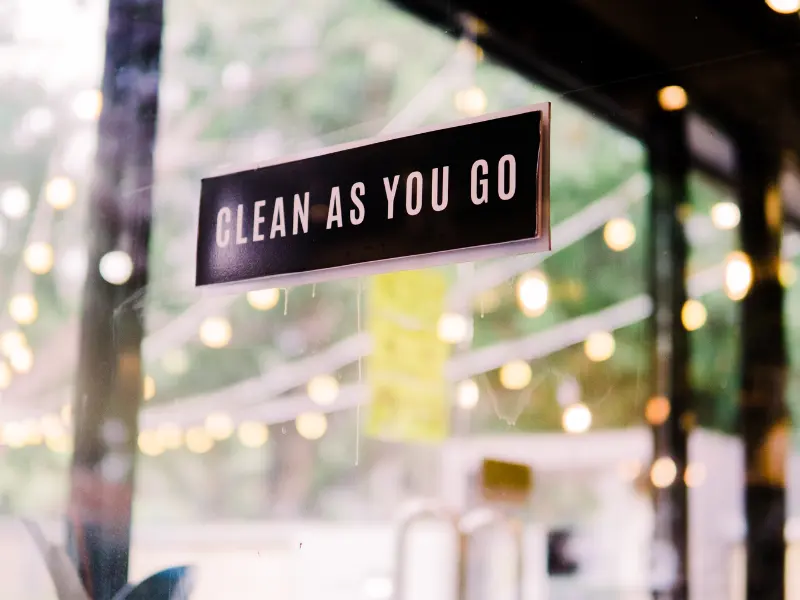 Sign reading "Clean as You Go" on a glass window.