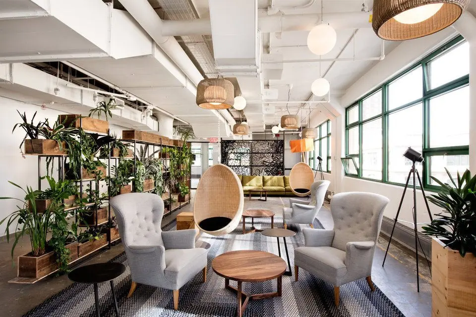 Modern lounge with gray chairs, hanging wicker chairs, wooden tables, and abundant plants. Large windows bring natural light. Relaxing vibe.